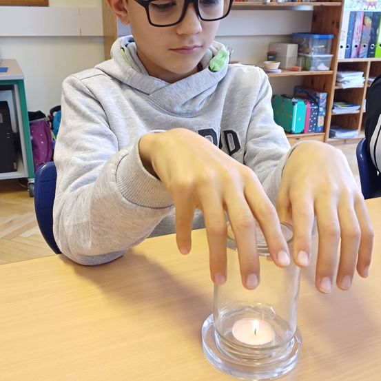 Ein junger Junge in einer grauen Kapuzenjacke sitzt an einem Tisch und hält eine Kerze in einem Glasbecher. Er scheint konzentriert zu sein, möglicherweise an einem Experiment oder einer Aktivität beteiligt. Hinter ihm steht ein Bücherregal mit verschiedenen Gegenständen.