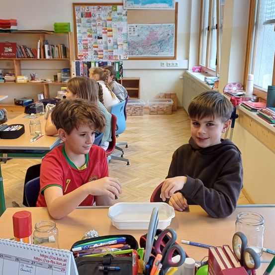 Zwei Jungen sitzen an einem Tisch in einem Klassenzimmer mit verschiedenen Schulmaterialien. Hinter ihnen sitzen andere Schüler an Tischen. Es gibt Regale mit Büchern und eine Pinnwand mit Bildern und Text.
