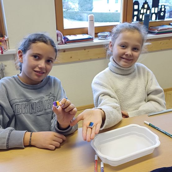 Zwei junge Mädchen sitzen an einem Tisch, lächeln und halten bunte Objekte. Das Mädchen auf der linken Seite trägt ein graues Sweatshirt. Das Mädchen auf der rechten Seite trägt ein weißes Rollkragenshirt. Ein durchsichtiger Behälter, Stifte und ein Fenster mit Blick auf Gebäude sind ebenfalls vorhanden.