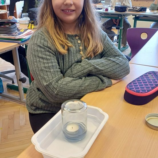 Ein junges Mädchen mit blonden Haaren sitzt lächelnd in einem Klassenzimmer. Ein Kerze in einem Glas steht auf ihrem Schreibtisch.
