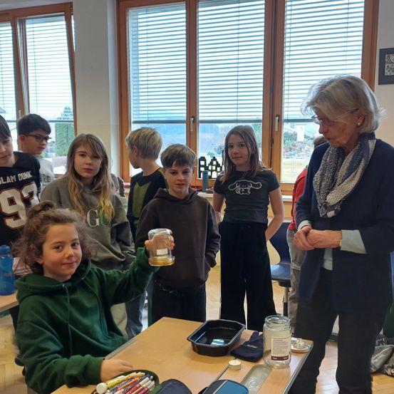 Eine Gruppe von Kindern in einem Klassenzimmer beobachtet eine Frau, die einen Glasbehälter mit einer Flüssigkeit darin hält. Sie stehen um einen Tisch mit verschiedenen Gegenständen. Der Raum hat große Fenster mit Jalousien.
