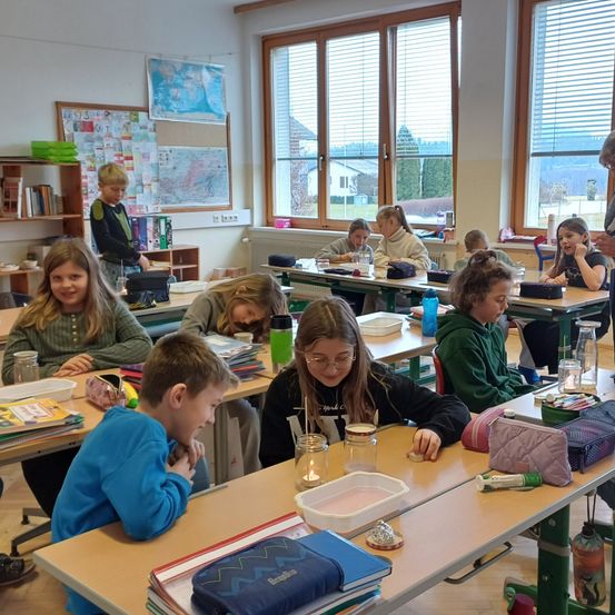Ein Klassenzimmer mit Schülern an Schreibtischen, ein Lehrer steht in der Nähe des Fensters. Die Schreibtische haben Bücher, Gläser und eine Kerze. Eine Karte und Bücher hängen an der Wand.