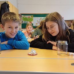 Drei Kinder in einem Klassenzimmer. Ein Junge in Blau lächelt in die Kamera, ein Mädchen in Schwarz lehnt sich mit den Armen auf den Tisch, und ein weiteres Mädchen in Rosa sitzt hinter ihnen. Ein Glasglas und eine kleine Kerze sind auf dem Tisch.