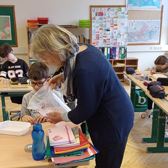 Eine Frau mit Brille hilft einem jungen Jungen mit Brille in einem Klassenzimmer mit anderen Schülern. Sie hält eine Plastiktüte mit Schulsachen. Es gibt Bücher in Regalen und Karten an der Wand.