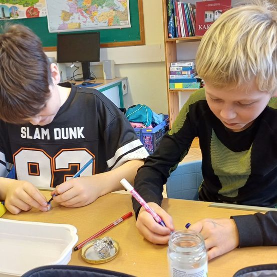 Zwei Jungen sitzen mit Zeichenmaterial am Tisch. Einer trägt ein schwarzes Slam Dunk-Trikot, der andere ein grünes und schwarzes Jackett. Sie zeichnen. Ein Regal mit Büchern steht hinter ihnen.