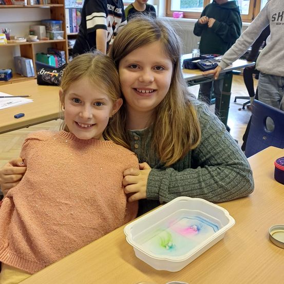 Zwei junge Mädchen lächeln, während sie an einem Tisch sitzen. Sie haben ihre Arme umeinander gelegt. Der Tisch hat einen Plastikbehälter mit farbiger Flüssigkeit darin.