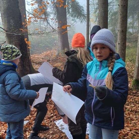 Eine Gruppe von Kindern, die für kaltes Wetter gekleidet sind, befindet sich in einem Wald. Sie halten Papiere in der Hand und scheinen etwas zu beobachten. Der Boden ist mit Blättern bedeckt.
