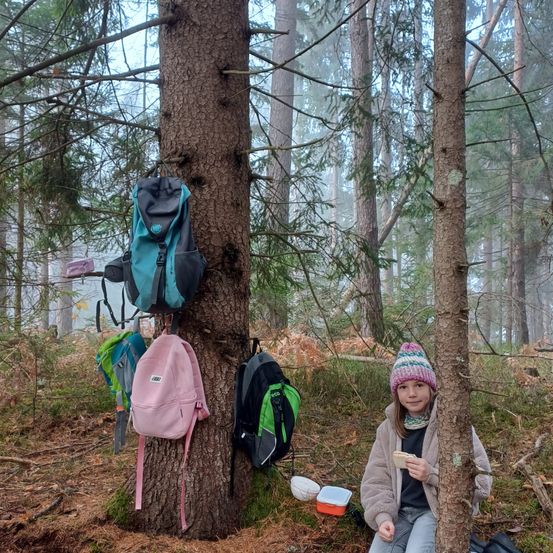 Ein junges Mädchen mit Mütze sitzt in einem Wald an einem Baum, mit mehreren Rucksäcken, die am Baum hängen, und einer Brotdose auf dem Boden.