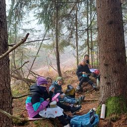 Fünf Personen sitzen auf dem Boden in einem Wald, einige in Winterkleidung. Sie essen und eine Person benutzt ein Handy. Ein Baumstamm ist neben ihnen.