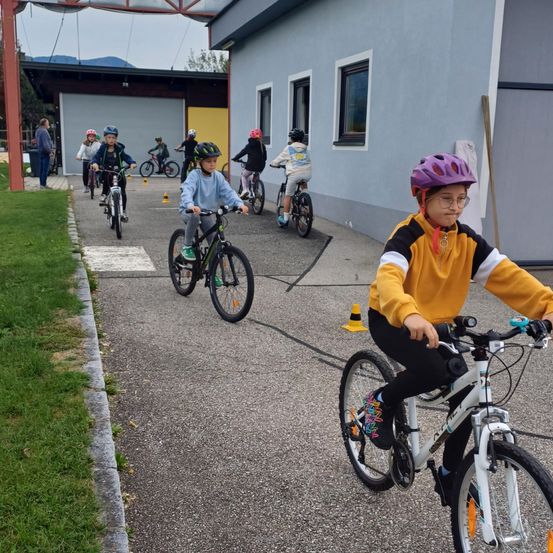 Mehrere Kinder, die Helme tragen, fahren auf einem asphaltierten Weg mit Fahrrädern. Sie navigieren durch auf dem Boden platzierte Kegel. Dahinter ist ein Gebäude mit einer gelben Tür und einer Garage zu sehen.