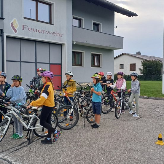 Eine Gruppe von Kindern steht mit ihren Fahrrädern vor einem Gebäude mit dem Wort Feuerwehr darauf. Die Kinder tragen Helme und Turnschuhe. Einige fahren ihre Fahrräder, während andere herumstehen. Es gibt mehrere Verkehrskegel auf der Straße.