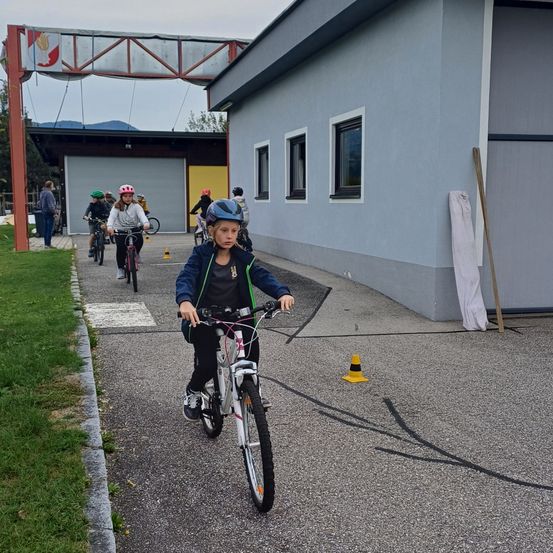 Kinder fahren auf Fahrrädern um ein Gebäude herum, wobei eine Person vorne fährt. Einige andere folgen hinterher. Das Gebäude hat eine gelbe Tür und graue Wände. Ein Mann geht in der Nähe.