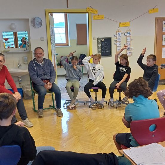 Eine Gruppe von Kindern und Erwachsenen sitzt auf Hockern in einem Klassenzimmer. Einige haben die Hände erhoben. Der Raum hat Holzböden und ein Waschbecken. Papiere und ein Spiegel sind im Hintergrund sichtbar.