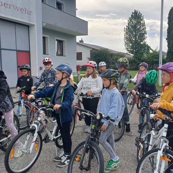 Eine Gruppe von Kindern mit Fahrrädern und Helmen steht vor einem Gebäude mit dem Wort Wehrwehr darauf. Die Kinder warten wahrscheinlich auf etwas.