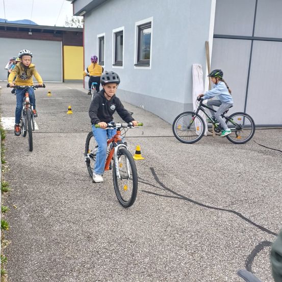 Mehrere Kinder fahren mit Fahrrädern vor einem Gebäude auf einem Schotterweg um Kegel.