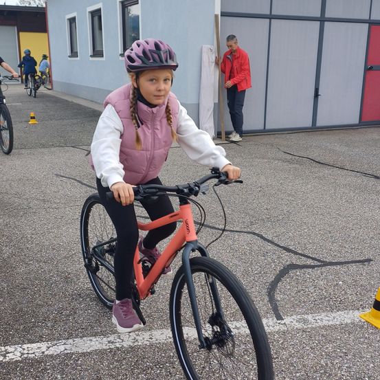 Ein junges Mädchen fährt auf einer gepflasterten Oberfläche mit einem Fahrrad. Sie trägt einen pinken Helm und eine Jacke. Ein Gebäude mit einer roten Tür ist im Hintergrund.