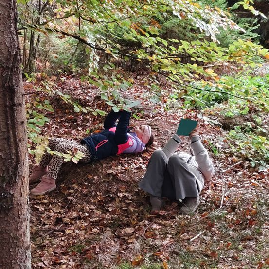 Zwei Personen liegen auf dem Boden im Wald, eine liest ein Buch, während die andere die Umgebung zu beobachten scheint.
