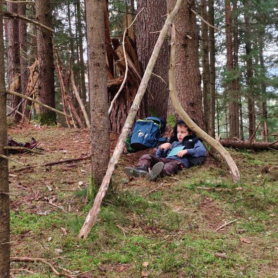 Ein junger Junge sitzt mit einem Rucksack und einem Buch auf dem Boden in einem Wald.