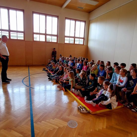 Ein Mann in einem weißen Shirt steht vor einer Gruppe von Kindern, die auf dem Boden in einer Turnhalle sitzen. Im Hintergrund befinden sich Fenster und Holzwände.