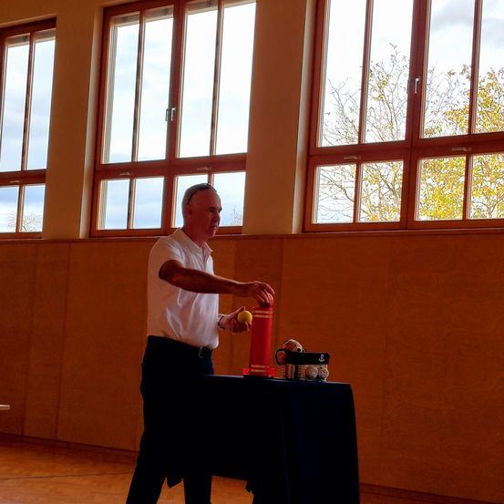 Ein Mann in einem weißen Hemd steht vor einem Tisch mit einem roten Rohr, einem gelben Ball und einem Behälter mit Spielzeug in einem Raum mit großen Fenstern.