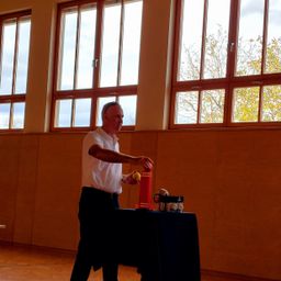 Ein Mann in einem weißen Hemd steht vor einem Tisch mit einem roten Rohr, einem gelben Ball und einem Behälter mit Spielzeug in einem Raum mit großen Fenstern.