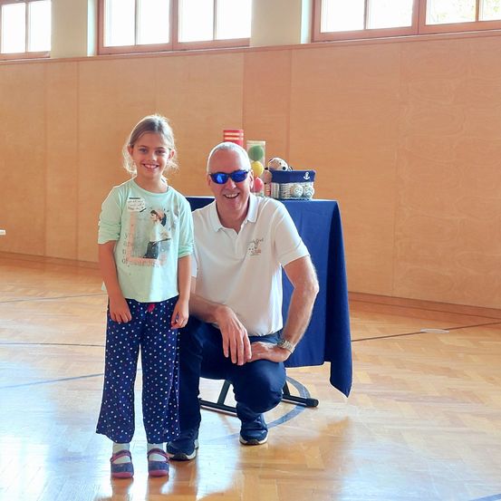 Ein Mann mit Sonnenbrille und Uhr kniet neben einem lächelnden Mädchen in einem Raum mit Holzböden und einem blauen Tisch.