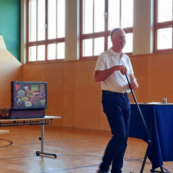 Ein Mann lächelt und hält einen Stock in der Hand, während er in einem Raum mit einem Koffer auf einem Tisch und einer blauen Tischdecke auf einem anderen Tisch geht.