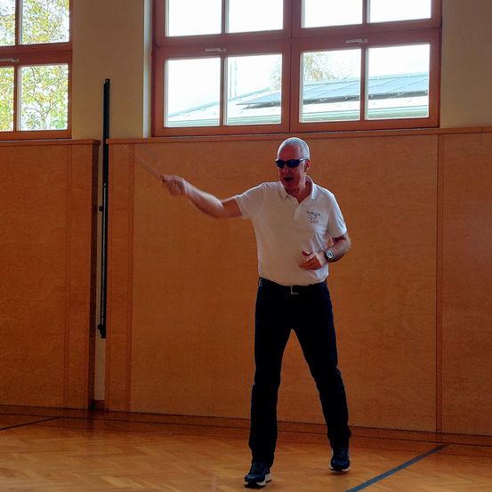 Ein älterer Mann in einer weißen Polo-Shirt und Sonnenbrille steht in einer Turnhalle mit einem Stock, wahrscheinlich bei einer Präsentation oder Anleitung.