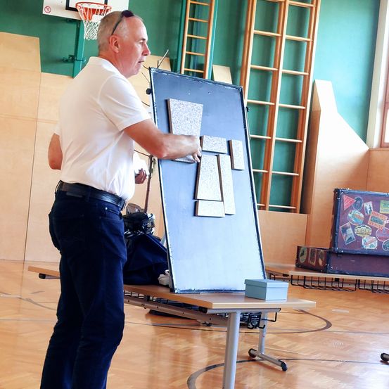 Ein älterer Mann steht in einer Turnhalle, hält ein blaues Brett mit Fliesen und einer Schachtel auf einem Tisch. Hinter ihm befinden sich Holzkonstruktionen, ein Basketballkorb und ein Fenster.