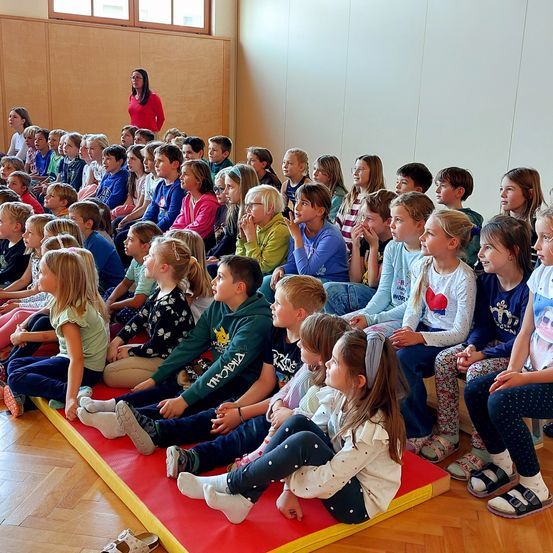 Ein Klassenzimmer voller Kinder, die aufmerksam eine Frau in einem roten Shirt beobachten, die vorne im Raum steht. Die Kinder sitzen auf gepolsterten Matten auf dem Boden.