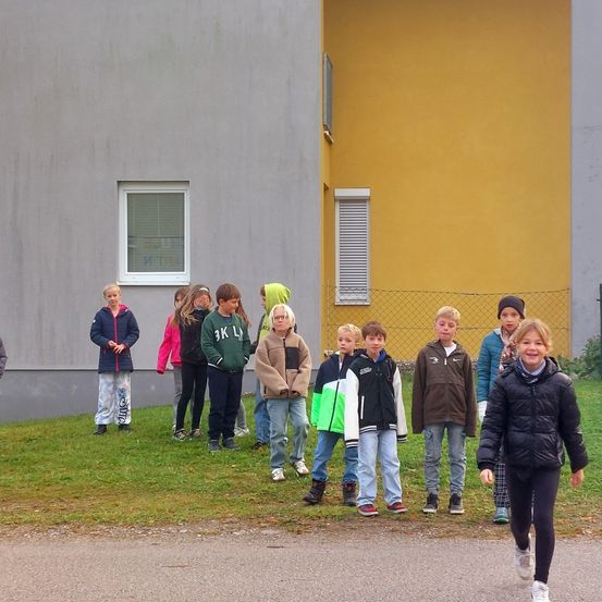 Eine Gruppe von Kindern steht auf dem Rasen vor einem gelben Gebäude. Sie tragen Jacken und Sneakers. Das Mädchen im Vordergrund lächelt.