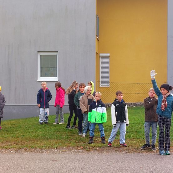 Eine Gruppe von Kindern in Winterkleidung steht auf einem Rasen vor einem Gebäude mit gelber Wand und Fenster. Ein Kind winkt.