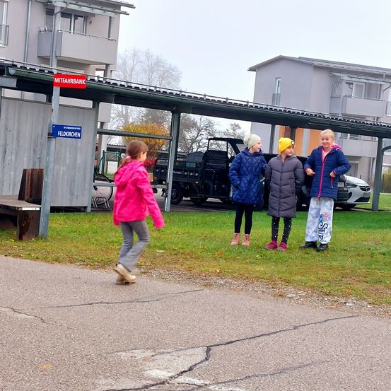 Vier Kinder stehen auf einem Parkplatz, zwei vor einer Bank und zwei vor geparkten Autos.
