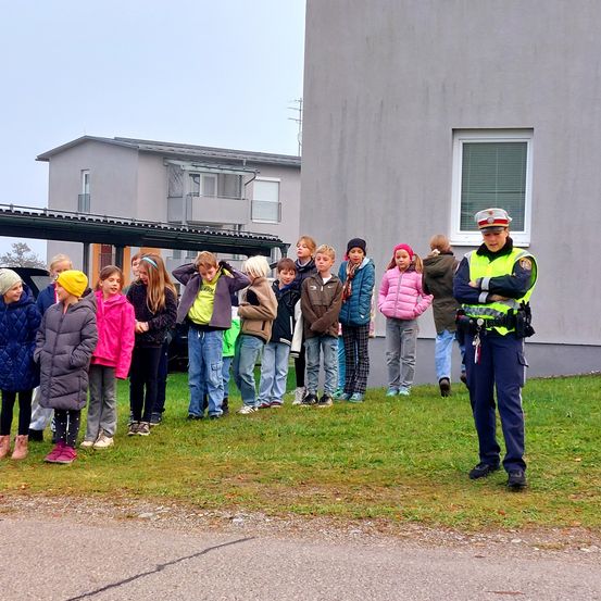 Eine Gruppe von Kindern, für das Wetter gekleidet, steht in einer Reihe mit einem Polizisten in einer reflektierenden Weste in der Nähe, vor einem Gebäude.