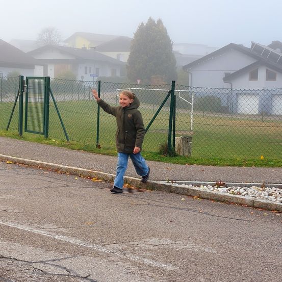 Ein junges Kind winkt in die Kamera, während es auf dem Bürgersteig neben einem Zaun und einem Fußballfeld steht.