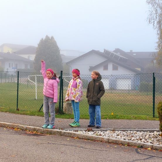 Drei Mädchen stehen auf einem Bürgersteig, eines mit erhobenem Arm. Sie tragen Jacken und Sneakers. Hinter ihnen ist ein Grasfeld von einem Zaun umschlossen. In der Ferne sind Häuser und Bäume von Nebel verhangen.