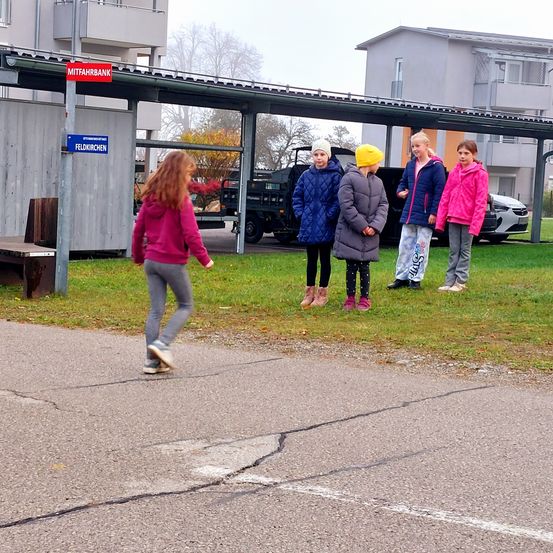 Mehrere junge Mädchen stehen auf einem Parkplatz, eine von ihnen geht. Ein Schild steht 'Mittagbank'. Im Hintergrund sind Gebäude und ein geparktes Auto zu sehen.