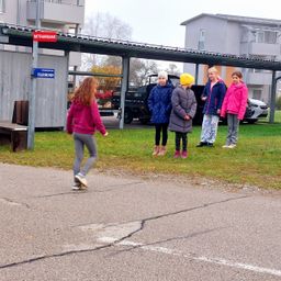 Mehrere junge Mädchen stehen auf einem Parkplatz, eine von ihnen geht. Ein Schild steht 'Mittagbank'. Im Hintergrund sind Gebäude und ein geparktes Auto zu sehen.