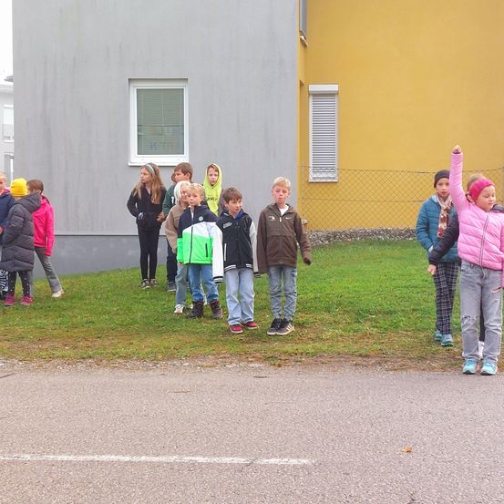 Eine Gruppe von Kindern steht auf einer Rasenfläche vor einem gelben Gebäude mit Fenstern und einem Zaun. Einige tragen Jacken, Mützen und Stiefel. Ein Kind hebt die Hand.