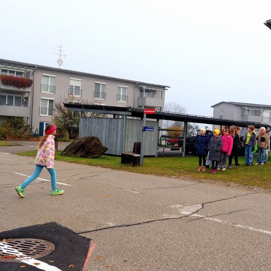 Ein junges Mädchen geht auf einem offenen Platz mit einem großen Felsen und einer Bank. Mehrere Kinder stehen auf einer Grasfläche in der Nähe eines Unterstands, und ein Gebäude befindet sich dahinter.