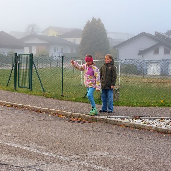 Zwei junge Mädchen stehen auf dem Bürgersteig, eine mit Stirnband und Turnschuhen, die andere in einer Jacke. Hinter ihnen trennt ein Zaun die Straße vom Rasen. Im Hintergrund sind mehrere Häuser in einer nebligen Atmosphäre zu sehen.