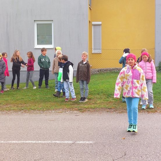Eine Gruppe von Kindern in Winterkleidung steht auf Gras neben einer Straße, mit Gebäuden im Hintergrund. Einige tragen Mützen und ein Kind hat Turnschuhe an.