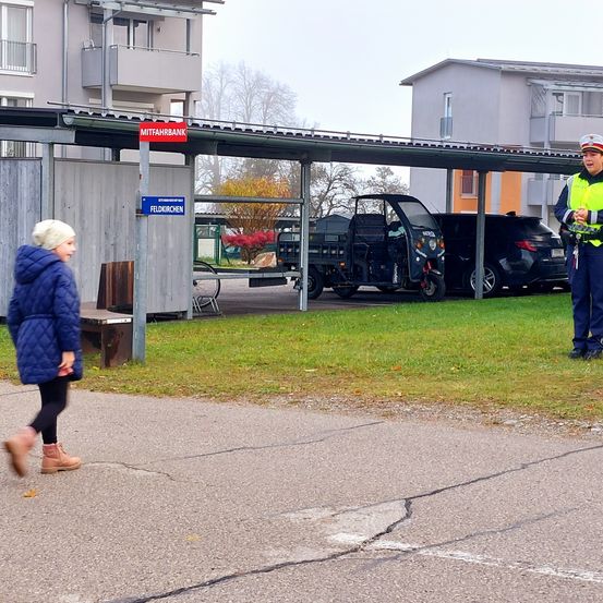 Eine Frau geht in der Nähe eines Parkplatzes mit einem Schild für 'Feldkirchen'. Ein Verkehrspolizist steht neben einem Fahrzeug. Im Hintergrund befinden sich Gebäude und Bäume.