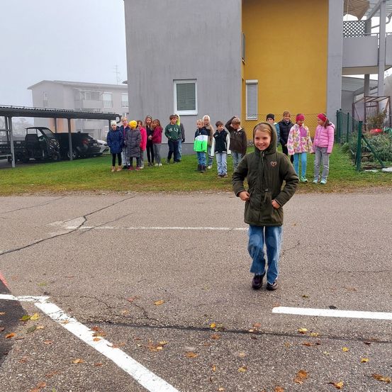 Ein kleines Mädchen steht lächelnd in der Mitte der Straße, während eine Gruppe von Kindern auf einem Rasenfeld in der Nähe eines gelben Gebäudes steht.