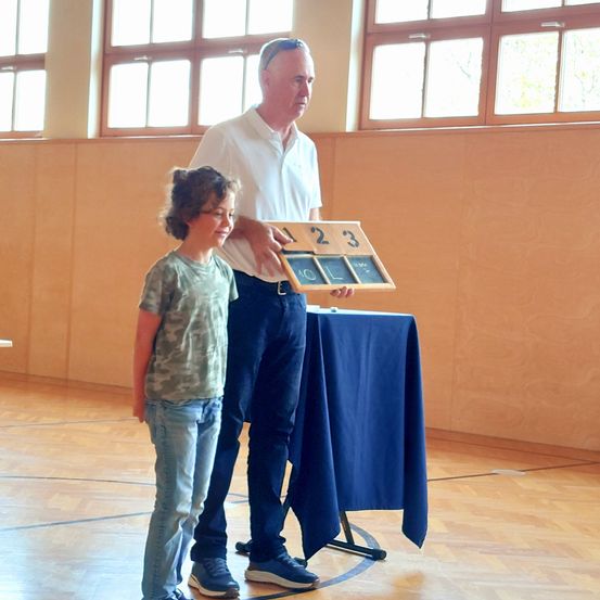 Ein älterer Mann hält ein Holznummernbrett, während ein junger Junge neben ihm in einer Turnhalle mit blauen Tischen und großen Fenstern steht.