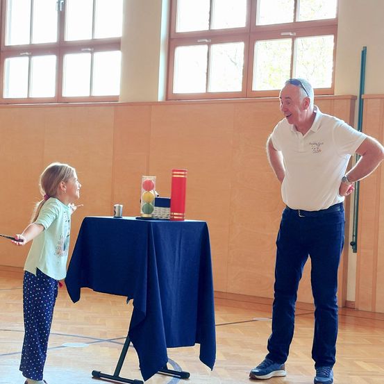 Ein Mann und ein kleines Mädchen sind drinnen. Der Mann lächelt und das Mädchen zeigt auf einen kleinen Tisch mit einem roten und weißen Gegenstand darauf. Das Mädchen trägt ein grünes Oberteil und blaue Hose. Der Mann trägt ein weißes Hemd, Jeans und Turnschuhe. Hinter ihnen ist eine Holzwand mit Fenstern.