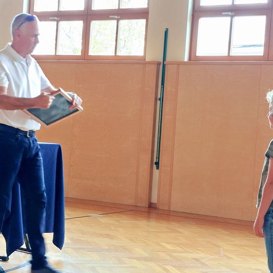 Ein Mann in einem weißen Hemd und Sonnenbrille hält eine Tafel in einem Raum mit Holzwänden und großen Fenstern. Ein Kind in Jeans steht rechts von ihm und schaut zu.