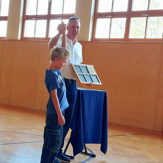 Ein älterer Mann gibt einem Jungen einen High-Five und hält eine Trophäe. Sie stehen in einem Raum mit Holzwänden und Fenstern.