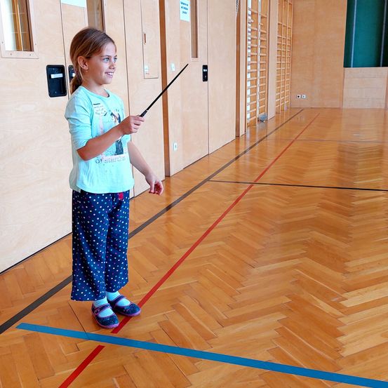 Ein kleines Mädchen steht in einer Turnhalle, hält einen Zauberstab und lächelt. Sie trägt ein blaues T-Shirt und Punktehosen. Die Turnhalle hat Holzböden und rote und blaue Linien. Hinter ihr befinden sich Holzschränke und eine grüne Wand.