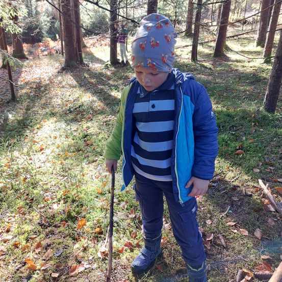 Ein kleiner Junge mit einem Fuchshut und gestreiftem Shirt, der einen Stock in einem Laubwald mit gefallenen Blättern hält.